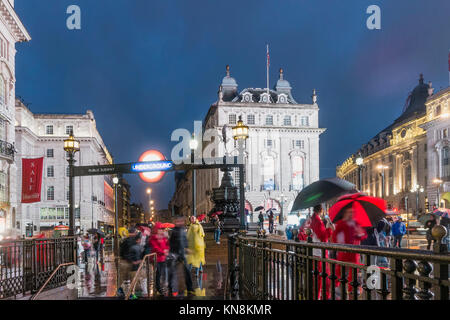 Persone con ombrelloni, Piccadilly Circus, pioggia, sera, ingresso della metropolitana di Londra, Regno Unito Foto Stock