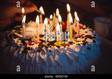 Torta di anniversario con la mano candele accese nel buio. Una mano con un fiammifero accende le candeline sulla torta di compleanno con crema bianca. 16 sedici candele o Foto Stock