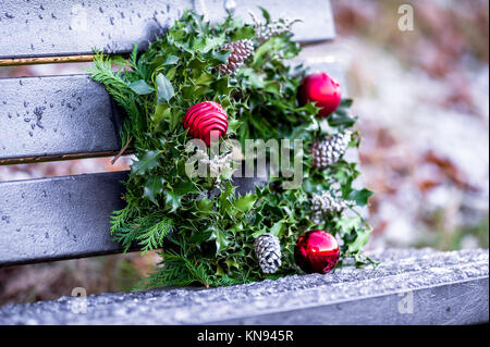 Decorativa ghirlanda di Natale fatte di pino e agrifoglio verde foglie e baubles rosso recante una panchina nel parco. Foto Stock