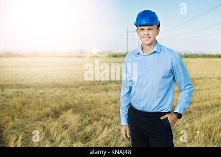 Da parte di un tecnico o di un lavoratore sorrisi nel casco protettivo in possesso di un telefono cellulare. Campi di grano, linea di alimentazione, giornata di sole e cielo blu. Foto Stock