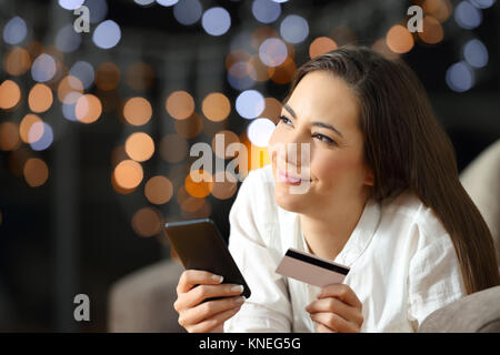 Ritratto di una donna felice chiedendo cosa comprare on line guardando il lato giacente su un divano nel soggiorno di casa nella notte Foto Stock