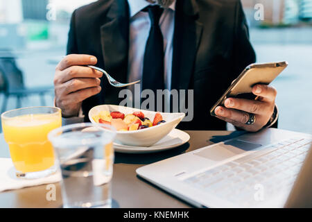 Imprenditore maturo seduti all'aperto, mangiare la colazione, utilizza lo smartphone, computer portatile sul tavolo, metà sezione Foto Stock