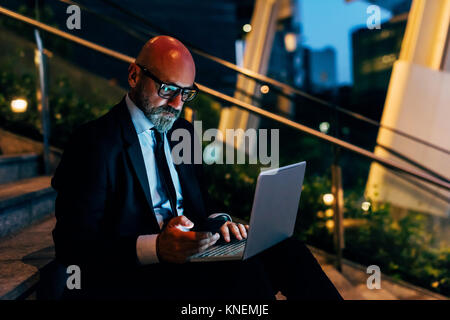 Imprenditore maturo all'aperto durante la notte, seduti su fasi, utilizzando laptop, smartphone di contenimento Foto Stock