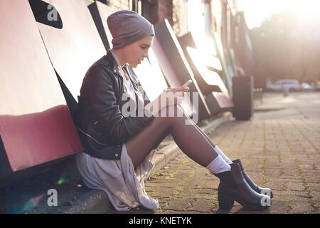 Giovane donna seduta su una strada di città guardando smartphone Foto Stock
