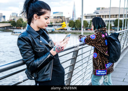 Giovane donna guardando smartphone con amico guardando fuori dalla passerella del millennio, London, Regno Unito Foto Stock