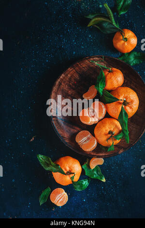 Agrumi su un piatto di legno con foglie verdi. La vibrante tangerini su uno sfondo scuro. Rustico fotografia di cibo. Foto Stock