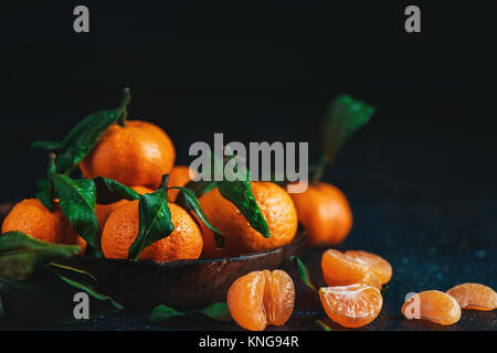Agrumi su un piatto di legno con foglie verdi. La vibrante tangerini su uno sfondo scuro. Rustico fotografia di cibo. Foto Stock