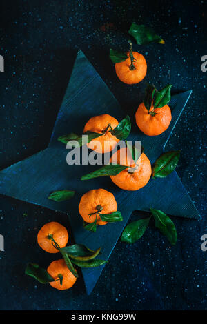 Orange tangerini con foglie verdi su una stella a forma di scheda di taglio. Frutti vibranti su uno sfondo scuro. Rustico fotografia di cibo. Foto Stock