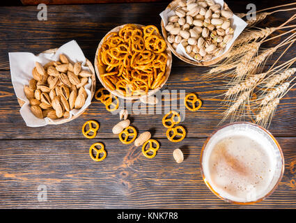 Vista superiore del bicchiere di birra con un grande capo di schiuma vicino a piastre con pistacchi, piccoli salatini e arachidi in legno scuro, scrivania. Cibo e bevande conce Foto Stock