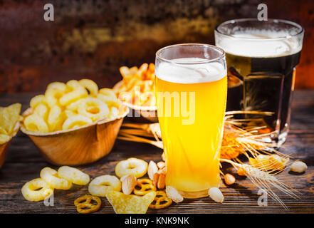 Due bicchieri con la luce filtrata e birra scura vicino a piastre con chips e snacks sparsi in legno scuro, scrivania. Cibo e bevande concept Foto Stock