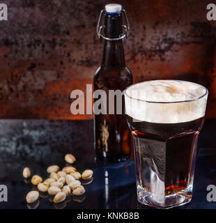 Vetro con appena spillata la birra scura, bottiglia di birra nei pressi di pistacchi sparsi sul nero di una superficie a specchio. Cibo e bevande concept Foto Stock