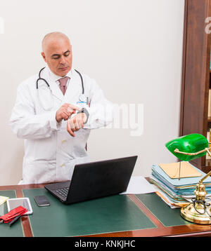 Sconcertato medico nel suo studio, utilizza un smartwacth davanti al suo computer portatile. Utilizzare le nuove tecnologie. Nel suo studio professionale, egli è seduto alla scrivania di antiquariato, un Foto Stock