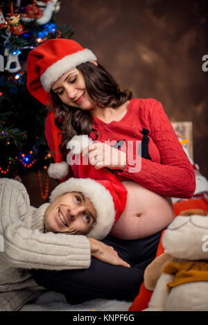 Close-up ritratto di famiglia del sorridente groom giacenti sulle gambe del suo amante incinta mentre lei sta giocando con la sua Santa Claus hat. Foto Stock