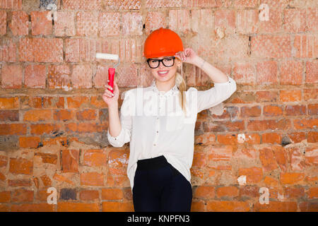 Ritratto di architetto studente o un pittore con rullo di vernice e proteggere il casco che indossa. Mattone sfondo rosso. Foto Stock