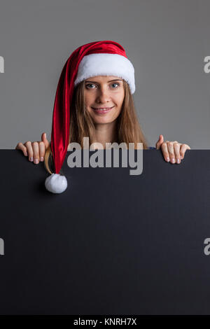 Donna che indossa santa natale hat holding e puntamento blank affissioni o segno targhetta copia spazio per la pubblicità di testo Foto Stock