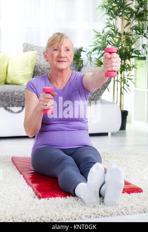 Senior donna seduta su un tappetino a casa e di esercitare con pesi Foto Stock