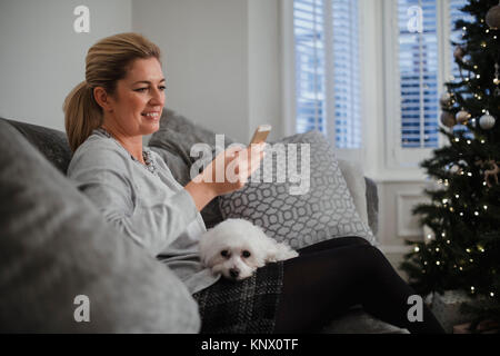 Donna matura è rilassante nel salotto di casa sua nel tempo di Natale. Il suo cane è seduta sul suo ginocchio e Lei sta usando un telefono intelligente. Foto Stock