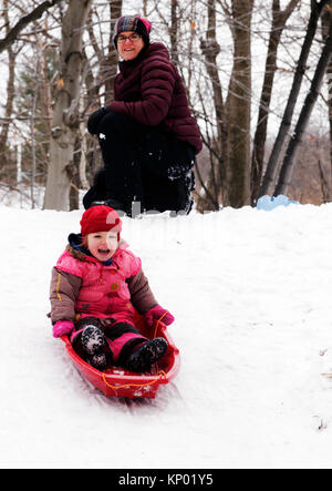 Una bambina (3 anni) in slittino Quebec, mentre la mamma si affaccia su Foto Stock