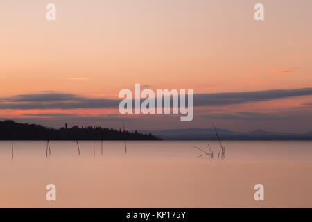 Una lunga esposizione vista del lago Trasimeno (Umbria, Italia) al tramonto, con poli e reti da pesca su perfettamente ancora acqua e un'isola in background Foto Stock