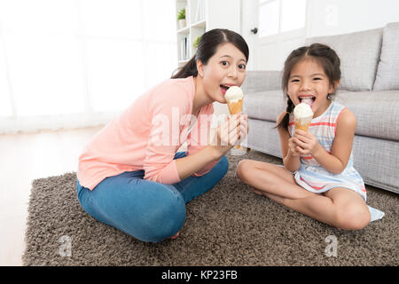 Allegro piacevole madre con bambina a mangiare il gelato insieme seduta sul soggiorno piano e di fronte alla telecamera che mostra godendo il concetto d'estate. Foto Stock