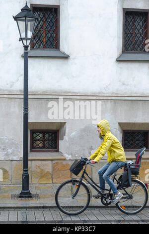 Pioggia femminile, veduta di una giovane donna che indossa un anorak giallo in bicicletta sotto la pioggia in via Grodzka, Cracovia, Polonia. Foto Stock