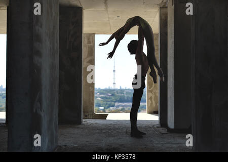 Silhouette di un paio di ballerini classici che posa sul pavimento in calcestruzzo dell'edificio incompiuto sul paesaggio urbano in background. Guy detiene una ragazza in th Foto Stock