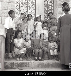 Degli anni Cinquanta, foto storiche, un barbuto sacerdote maschio con hat e accappatoio ha un po' di divertimento con un gruppo di bambini seduti e in piedi sui gradini al di fuori di un ingresso sincronizzato, Giordania. Foto Stock