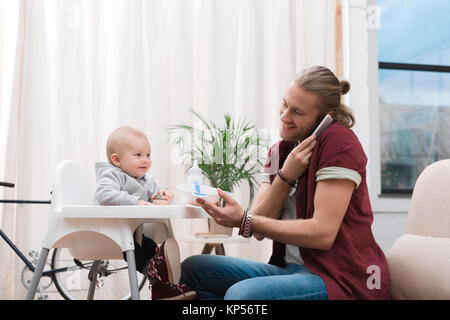 Padre alimentando il suo piccolo figlio con cibo per bambini mentre parla sullo smartphone Foto Stock