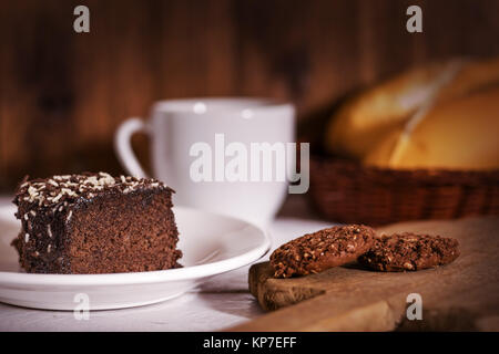 Torta al cioccolato, biscotti, caffè tazza di porcellana e pane polpettine sulla sommità del legno di un tavolo bianco superiore e uno sfondo di legno Foto Stock