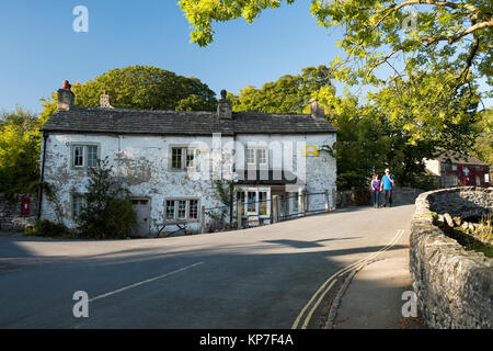 Sulla giornata di sole, giovane sono a piedi attraverso il ponte di pietra nel centro Malham, dal vecchio negozio del villaggio con peeling vernice bianca - Yorkshire Dales, Inghilterra, Regno Unito. Foto Stock