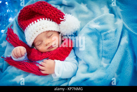 Ritratto di un piccolo grazioso baby boy indossando festosa Santa hat e dormono a casa, coperto con il suo blu coperta come nella neve, felice vacanze di Natale Foto Stock