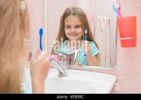 Sei anni di ragazza con uno spazzolino da denti e si guarda allo specchio, mentre nella stanza da bagno Foto Stock