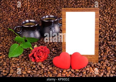 Cornice per foto con sfondo bianco, due cuori, una rosa rossa e due tazze di caffè nero su uno sfondo di chicchi di caffè. Foto Stock