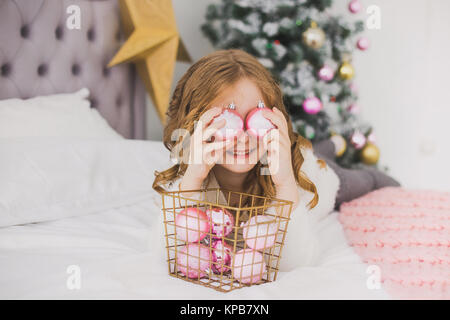 Ritratto di carino felice bambina la mattina di Natale in home interni. Bambino pagando con decorazioni di vacanza, rendendo occhio di sfere rosa mentre posa Foto Stock