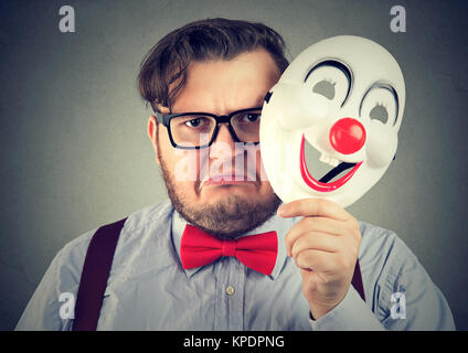 Giovane uomo arrabbiato accigliata mentre guarda la fotocamera e azienda happy clown maschera. Foto Stock