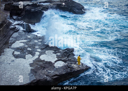Un pescatore solitario in piedi sul bordo di una roccia di fronte a una massiccia ondata di colpire la riva Foto Stock