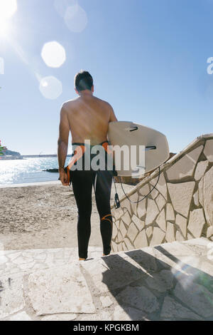 L'uomo sulla sua schiena, con la tavola da surf sotto il suo braccio e una muta umida, andare giù per una rampa di scale per arrivare alla spiaggia Foto Stock