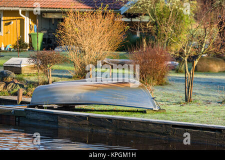 Piccolo in alluminio canotto capovolto sul prato lungo il fiume in un freddo gelido e la mattina. Casa Gialla e arbusti in background. Foto Stock