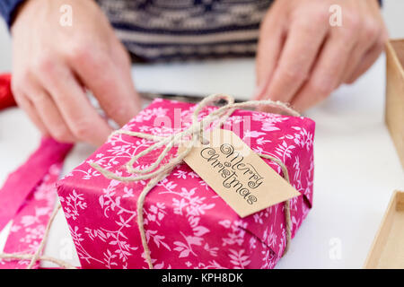 Primo piano di un giovane uomo caucasico legatura di un regalo con una stringa da cui pende una tag di swing con il testo buon natale Foto Stock