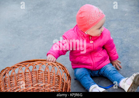 Un anno-vecchio figlio in una camicia rosa e hat siede sul calcestruzzo accanto a un cesto di vimini Foto Stock