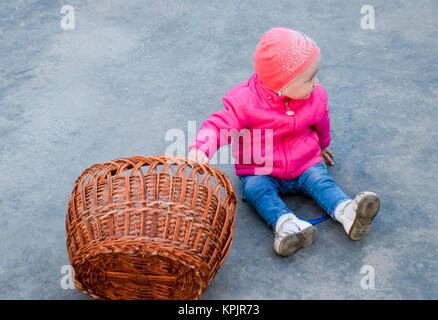 Un anno-vecchio figlio in una camicia rosa e hat siede sul calcestruzzo accanto a un cesto di vimini Foto Stock