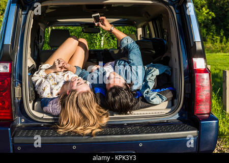 Coppia giovane che stabilisce nel retro del loro veicolo impaccato in un viaggio su strada che mostra i loro stivali e con il loro telefono cellulare; Edmonton, Alberta, Canada Foto Stock