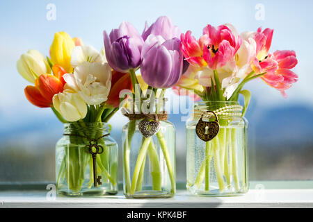 Varie tulipani colorati in vasi di vetro con ciondoli decorativi sul davanzale della finestra con la città dietro; Surrey, British Columbia, Canada Foto Stock