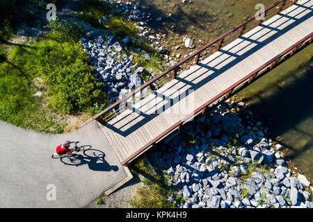 Vista da direttamente al di sopra di un ciclista in una maglietta rossa andando giù per un sentiero e su un ponte su un fiume; Calgary, Alberta, Canada Foto Stock