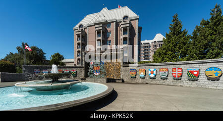 Colorato provinciale creste simbolico su un muro di pietra e fontana di acqua lungo un marciapiede; Victoria, British Columbia, Canada Foto Stock