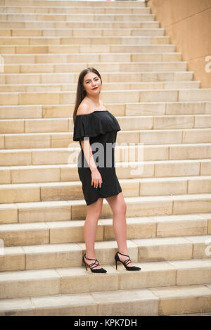 Bella donna felice indossando abito nero e tacchi alti e in piedi sui gradini esterni sorridente Foto Stock