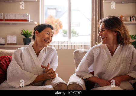 Due donne sorridente in accappatoio in seduta area ricevimento e parlare prima del trattamento termale. Due amiche rilassante nel centro di bellezza e benessere in sala d'attesa e s Foto Stock