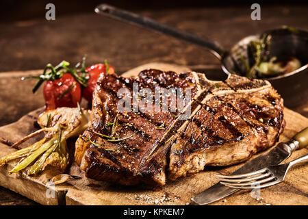 Alla griglia preparata in grandi t-bone steak circondato da pomodori e guarnita con condimenti accanto alla forchetta e coltello sul tagliere Foto Stock