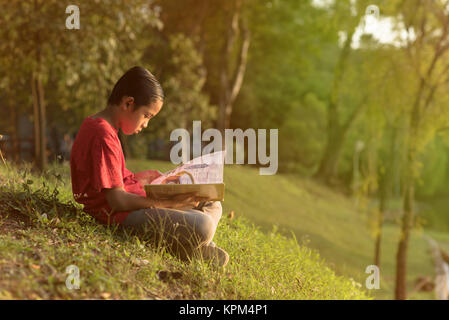 Giovane ragazzo asiatico in maglietta rossa per leggere un libro vicino al giardino del lago in serata Golden Sun ora Foto Stock