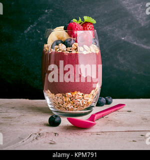 Deliziosa Acai berry e avena dessert con pezzi di banana, fragola e mirtillo in vetro di grandi dimensioni accanto al cucchiaio sul centro del tavolo in legno con sfondo scuro Foto Stock